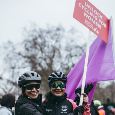 Two women of colour with black cycle helmets and shades hold up a red LCC placard that says Unlock Cycling For Women at protest